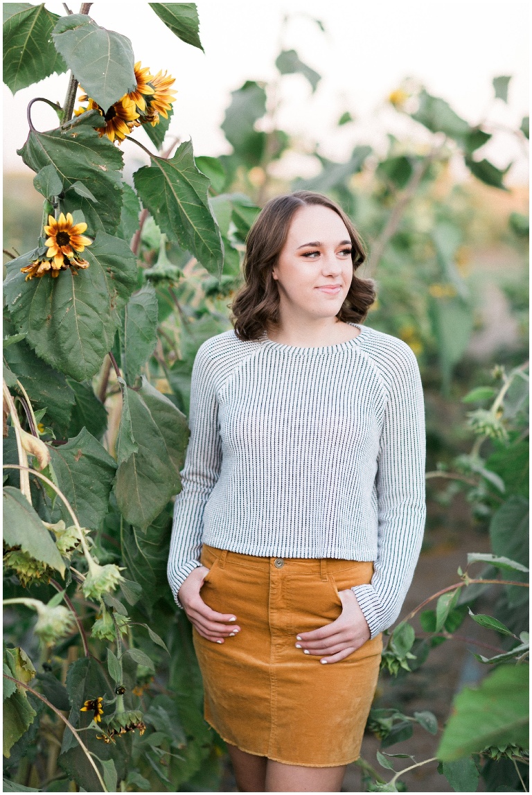 girl in a sweater and skirt standing next to sunflowers
