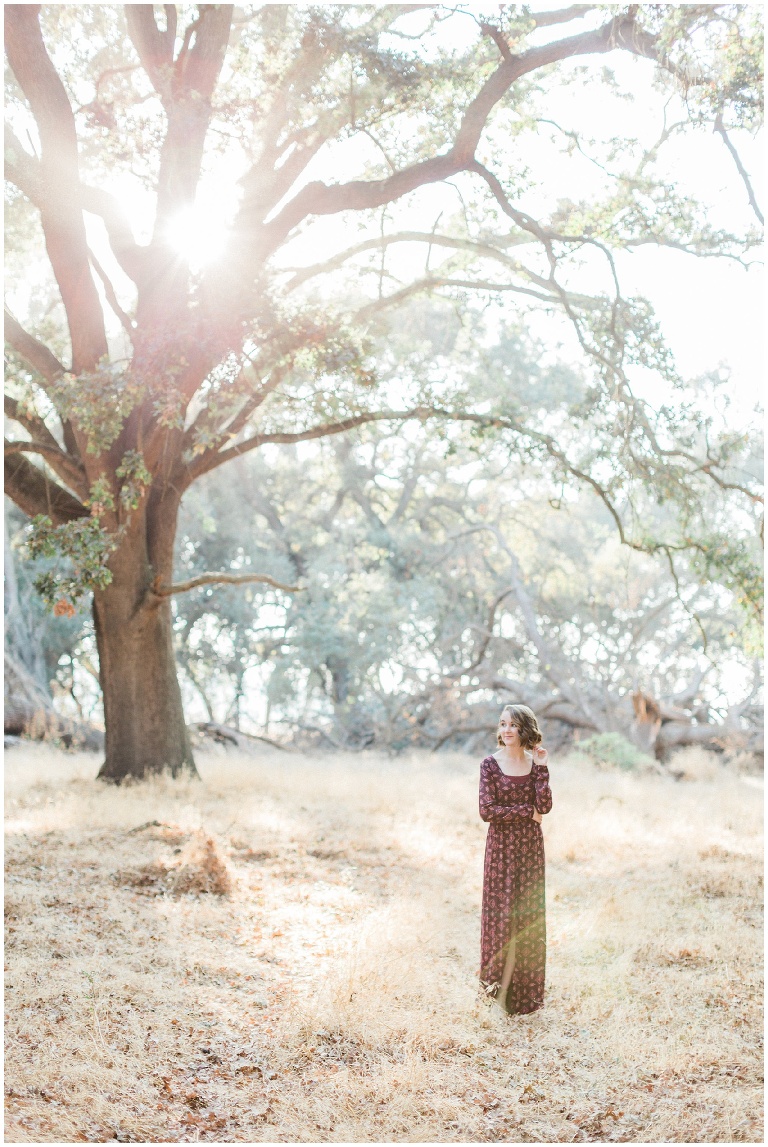 girl in a maroon dress standing under an oak tree with the sunlight coming through
