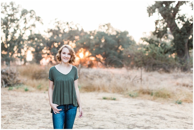 girl in a green shirt and jeans standing in front of the sunset in an open field