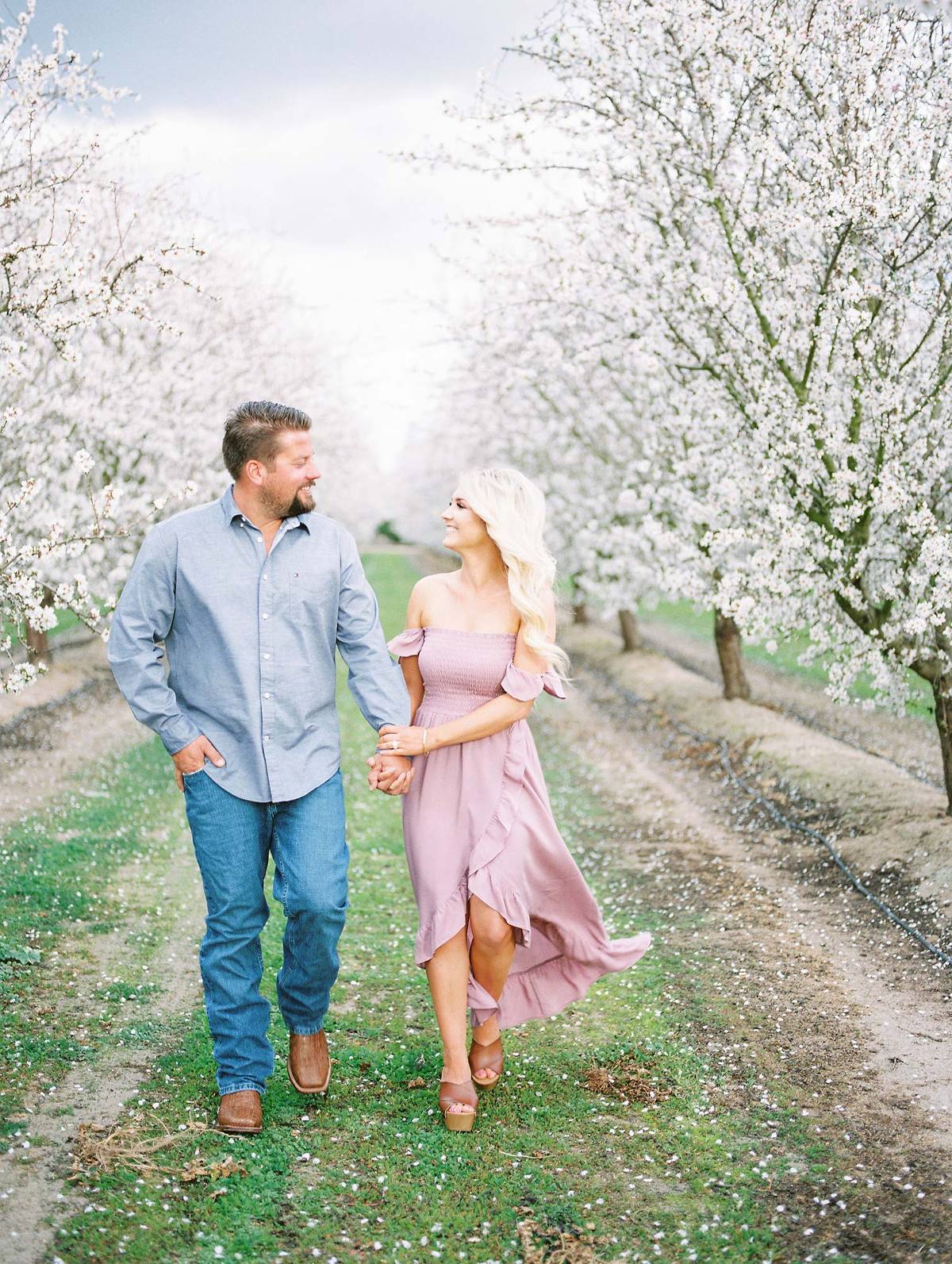 couple holding hands walking through an almond blossom orchard on a cloudy day